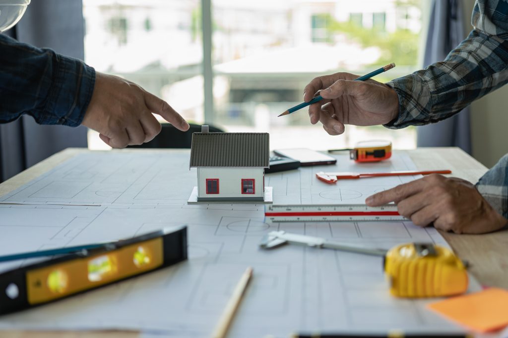 Zwei Personen besprechen ein Architekturmodell eines Hauses, das auf einem Bauplan platziert ist, umgeben von Messwerkzeugen wie einem Lineal, einer Wasserwaage und einem Maßband.