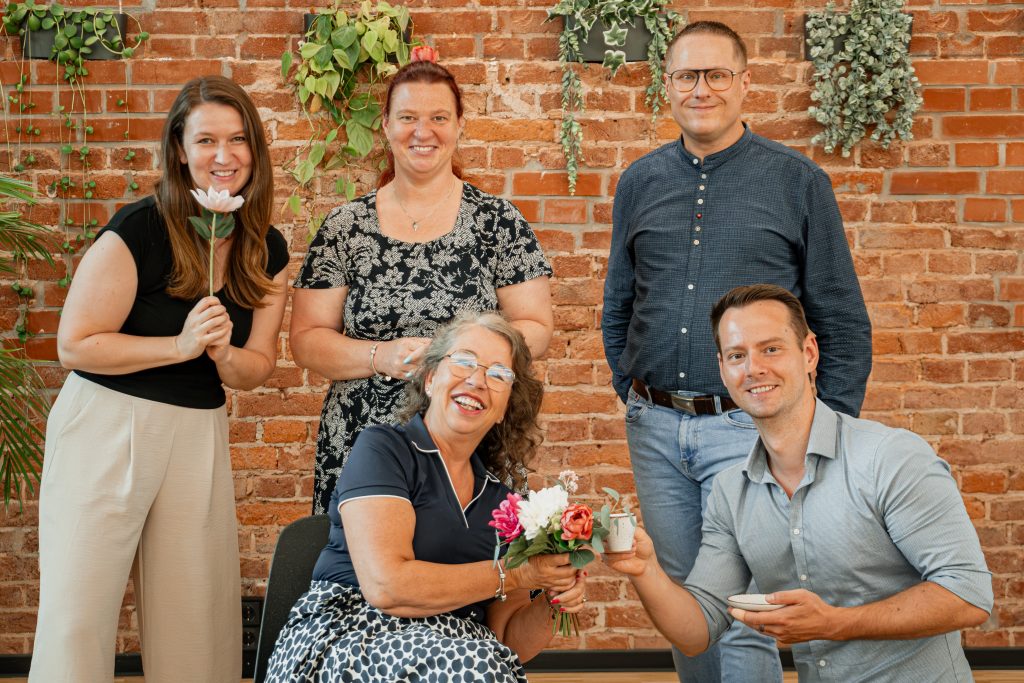 Entspanntes Gruppenfoto des SONAPRO-Beratungsteams vor einer rustikalen Backsteinwand mit grünen Hängepflanzen. Fünf gut gelaunte Personen posieren für das Bild: Im Vordergrund sitzt eine Frau mit Brille und lacht herzlich, während sie einen kleinen Blumenstrauß und eine Tasse hält. Eine weitere Frau kniet neben ihr und stößt mit einer kleineren Tasse an. Im Hintergrund stehen drei weitere Teammitglieder: eine Frau mit weißer Blume in der Hand, eine Frau in einem schwarz-musternden Kleid und ein Mann mit Brille, alle lächelnd in die Kamera blickend.