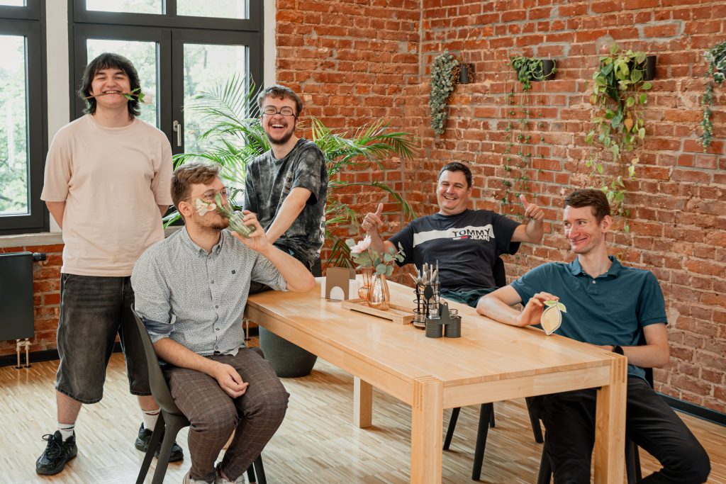Ein humorvolles Gruppenfoto des Entwicklungsteams an einem Holztisch vor einer rustikalen Backsteinwand. Die fünf Teammitglieder zeigen sich in einer ausgelassenen Stimmung: Ein Mitarbeiter steht links und hält eine Blume im Mund, ein zweiter sitzt und hält ein kleines Glas vor sein Gesicht, ein dritter steht dahinter und lacht herzlich, ein vierter sitzt am Tisch und zeigt mit beiden Händen Daumen hoch, und der fünfte sitzt rechts am Tisch und hält eine Zitronen-Grafik in die Kamera.