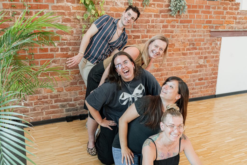Ein humorvolles Gruppenfoto des Marketing- und Vertriebsteams vor einer rustikalen Backsteinwand. Die fünf Teammitglieder haben sich in einer spielerischen Formation hintereinander aufgestellt, wobei sie sich gegenseitig über die Schultern lehnen. Alle Personen lachen oder blicken fröhlich in die Kamera, was eine ausgelassene und dynamische Atmosphäre im Team widerspiegelt.