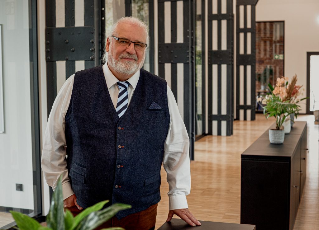 Porträt von Jens Knöfler, geschäftsführender Gesellschafter. Er steht selbstbewusst in einem modernen Büro mit architektonisch markanten schwarz-weiß gestreiften Säulen. Er trägt ein weißes Hemd mit einer gestreiften Krawatte und eine dunkelblaue Weste. Sein Blick ist freundlich und direkt in die Kamera gerichtet.