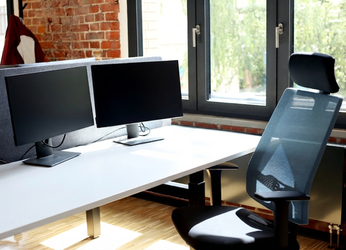 Moderner und heller Arbeitsplatz im SONAPRO Büro: Die Aufnahme zeigt einen ergonomisch eingerichteten Schreibtisch mit zwei Bildschirmen und einem modernen Bürostuhl in einer ansprechenden Arbeitsumgebung mit Ziegelsteinwand.